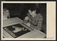 [recto] A young artist of Japanese parentage, correcting a poster in the Poster Shop at the Heart Mountain Relocation Center. Posters, ...