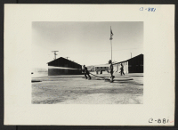 [recto] Manzanar, Calif.--Fire equipment is used in spraying the ground to keep the dust down at this War Relocation Authority center. Administration buildings in background. ;  Photographer: Lange, Dorothea ;  Manzanar, California.