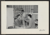 [recto] A patient, teacher of high school science, being examined in a temporary field clinic by Doctor K. H. Taira. In the background Pharmacist Tom Arase prepares a prescription. (L to R) Patient; Tom Arase, Pharmacist; Kitti Tairce [?], M.D. ;  Photographer: