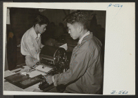 [recto] Editor Albert Saijo inspects a copy of Echo, high school paper, being run off on the mimeograph machine by Toyoji Sugita, staff artist and technician. ;  Photographer: Hosokawa, Bill ;  Heart Mountain, Wyoming.