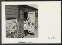 [recto] A mother and daughter, Issei and Nisei, who have been living in this Assembly Center for one month, are seen at the door of their home in the barracks. ;  Photographer: Lange, Dorothea ;  San Bruno, California.