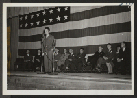 [recto] Sgt. Kuroki delivers an address in the high school auditorium. In the background are community councilmen, Project Director Robertson, Nobu Kawai (associate editor of the Sentinel), Hisa Hirashiki of Community activities, and Geo. Nakaki, Councilman. ;