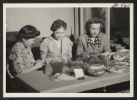 [recto] These three women, two of them Issei resettlers, are assembling artificial flower novelties in a workroom of the Miya Flower ...