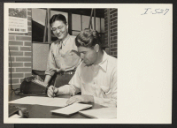 [recto] Mr. B. J. Wilson, Industrial Engineer, and George Okamura, an evacuee from Poston, Arizona, are shown discussing their office problems ...