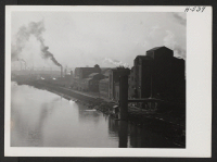 [recto] That Peoria, Illinois, has many thriving industries is easily seen as one enters the city from the east going over the Illinois River, along which border many factories such as the kind seen in this picture, which looks south along the west bank of the ri
