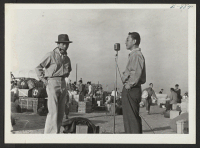 [recto] Toshio Ninomiya, left, assisted with registration and Tomoo Ogita, right, at the microphone giving instructions to new residents. ;  Photographer: McClelland, Joe ;  Amache, Colorado.