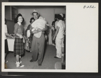 [recto] Having finished with the brief medical inspection, new arrivals at Topaz from the Tule Lake Center are shown being directed to the house committee. A father is shown carrying one of the many infants that arrived on trip 14. ;  Photographer: Mace, Charle