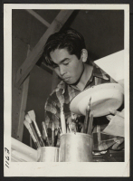 [recto] A young poster artist intent on applying color for a poster, in the [illegible] Poster Shop at the Heart Mountain Relocation Center. The Poster Shop provides posted notices for center activities and safety. ;  Photographer: Parker, Tom ;  Heart Mounta