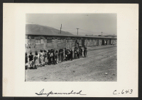 [recto] View of the Tanforan Assembly Center, showing a type of barrack peculiar to the center. Also shown is part of a line-up of evacuees waiting for places in the mess hall for their noon meal. ;  Photographer: Lange, Dorothea ;  San Bruno, California.