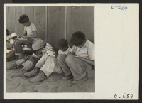 [recto] Manzanar, Calif.--Young sixth-grade students studying their lessons in the shade of the barracks at this first voluntary elementary school. ;  Photographer: Lange, Dorothea ;  Manzanar, California.