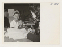 [recto] With her son, young Dennis, in her arms, Mrs. T. Sasabushi, Nisei resident at the Granada center, registers for defense ...