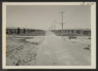 [recto] Looking down a street of the Minidoka Relocation Center. A service flag with two stars can be seen in the window of the first room in the barrack at extreme right. ;  Hunt, Idaho.