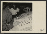 [recto] A young artist in the poster shop at the Heart Mountain Relocation Center copies a poster in Japanese for the ...
