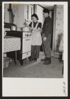 [recto] Over a Skelgas cooking range, Mrs. Bill Okazaki prepares the noonday meal for her husband, as he looks on. The ...