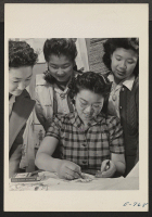 [recto] A closeup of textile work. Yasko Taniguchi (seated) is working on a textile with crayons. In the background watching her work are, left to right, Terumi Uyesugi, Sumiye Sasaki and Jane Nakamura. ;  Photographer: Coffey, Pat ;  Amache, Colorado.