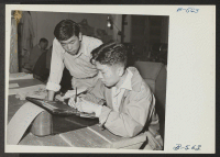 [recto] Editor Saijo watches while Toyoji Sugita, artist, puts finishing touches to a headline on the stencil used for mimeographing Echoes, the Heart Mountain high school student newspaper. ;  Photographer: Hosokawa, Bill ;  Heart Mountain, Wyoming.